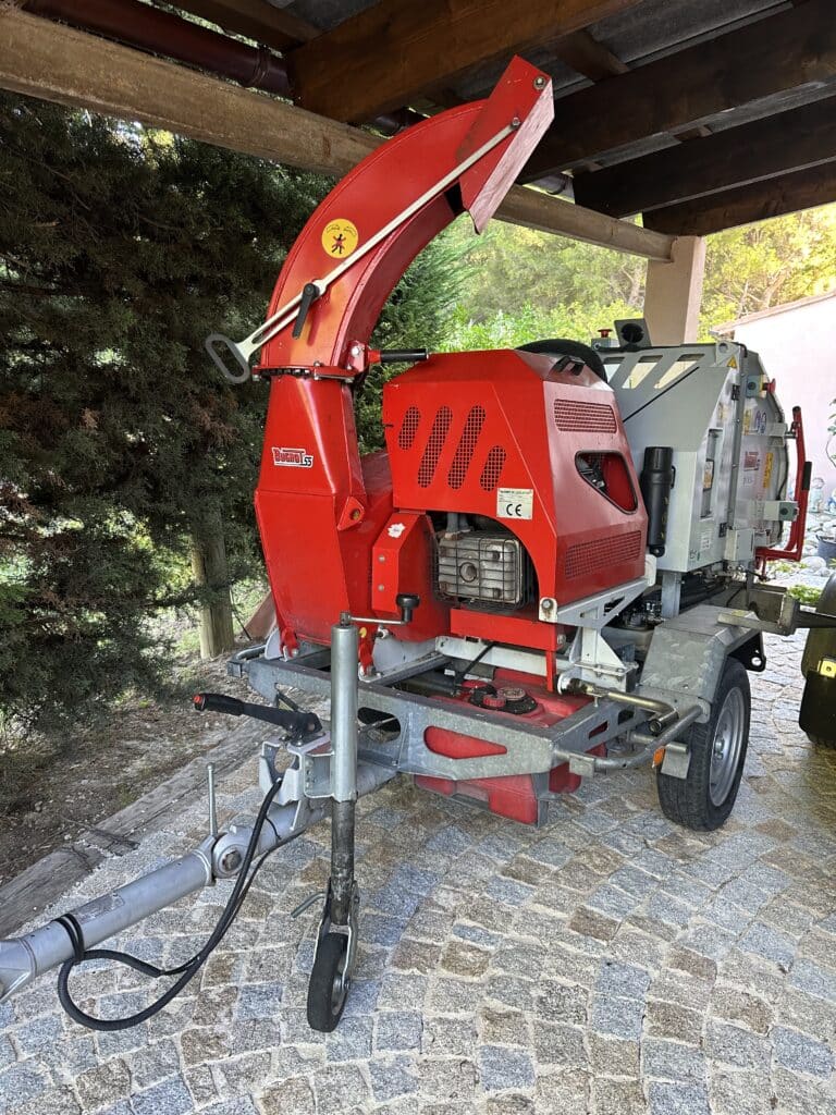 Broyeur de branches Bugnot 55 rouge et gris sur remorque, stationné sous un abri en bois sur pavés.