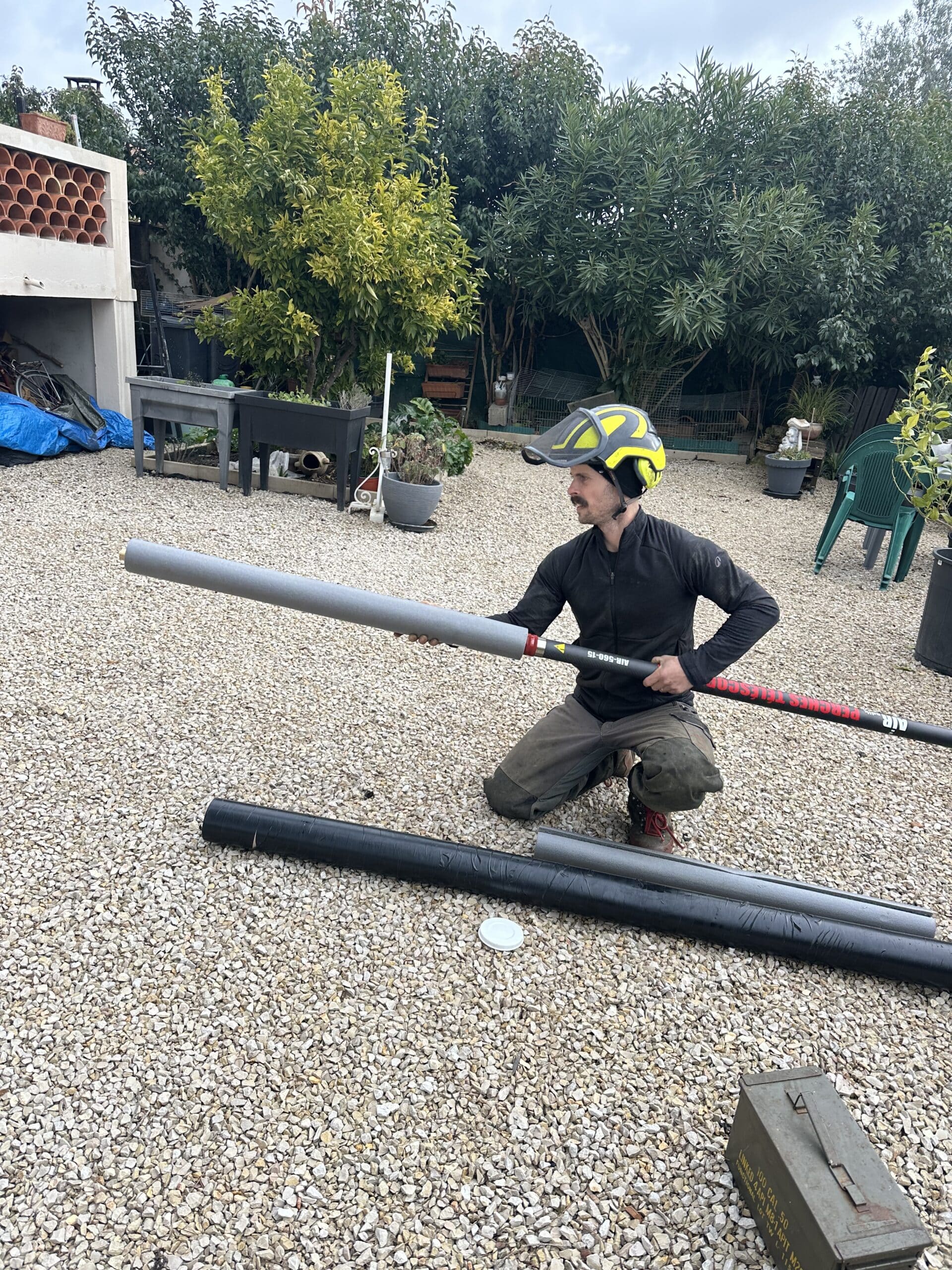 Homme en casque de sécurité jaune à genoux sur du gravier, assemblant une perche télescopique AIR-560-15. Arrière-plan de jardin.